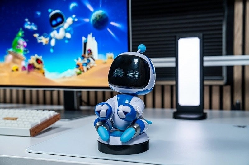 Бело-синяя фигурка-подставка Astro Bot на чёрном основании стоит на столе перед монитором с игрой