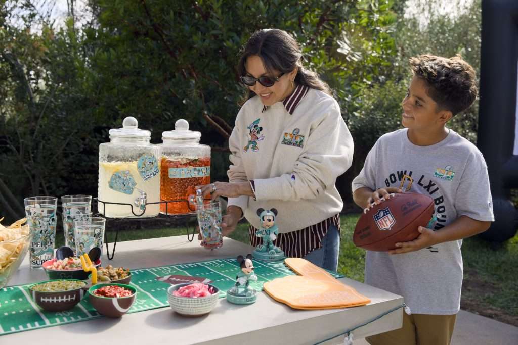 Промофото Disney × NFL: женщина и ребёнок в тематической одежде рядом со стаканами и фигурками Микки на столе