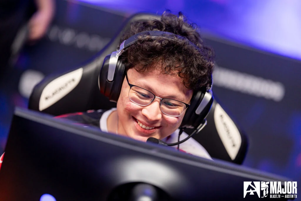 Игрок Nemiga khaN в очках и гарнитуре улыбается за монитором на турнире BLAST Austin Major