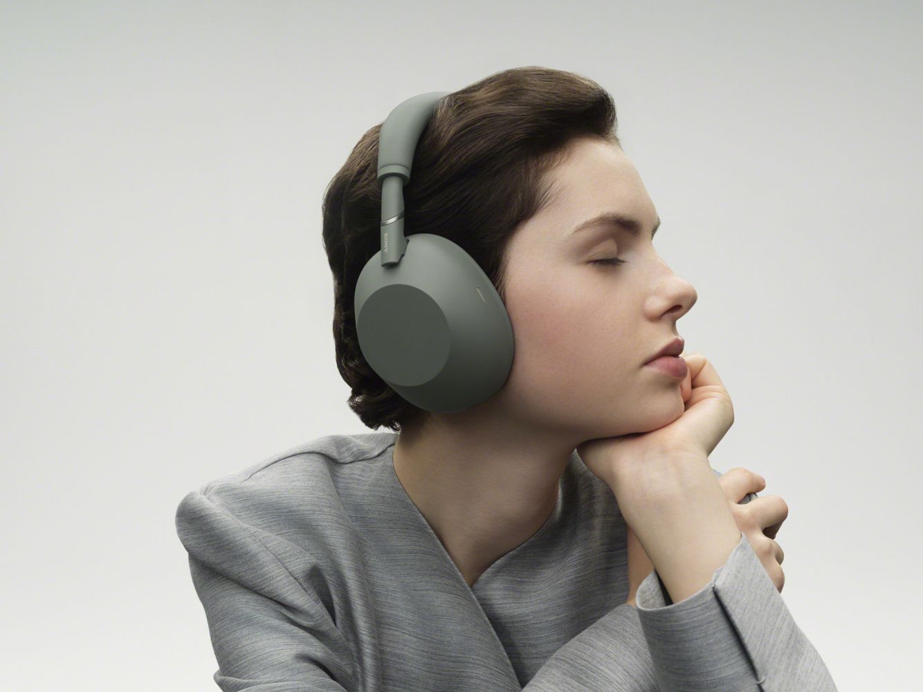 Человек в серой кофте слушает полноразмерные наушники Sony WH-1000XM6 оливково-серого цвета