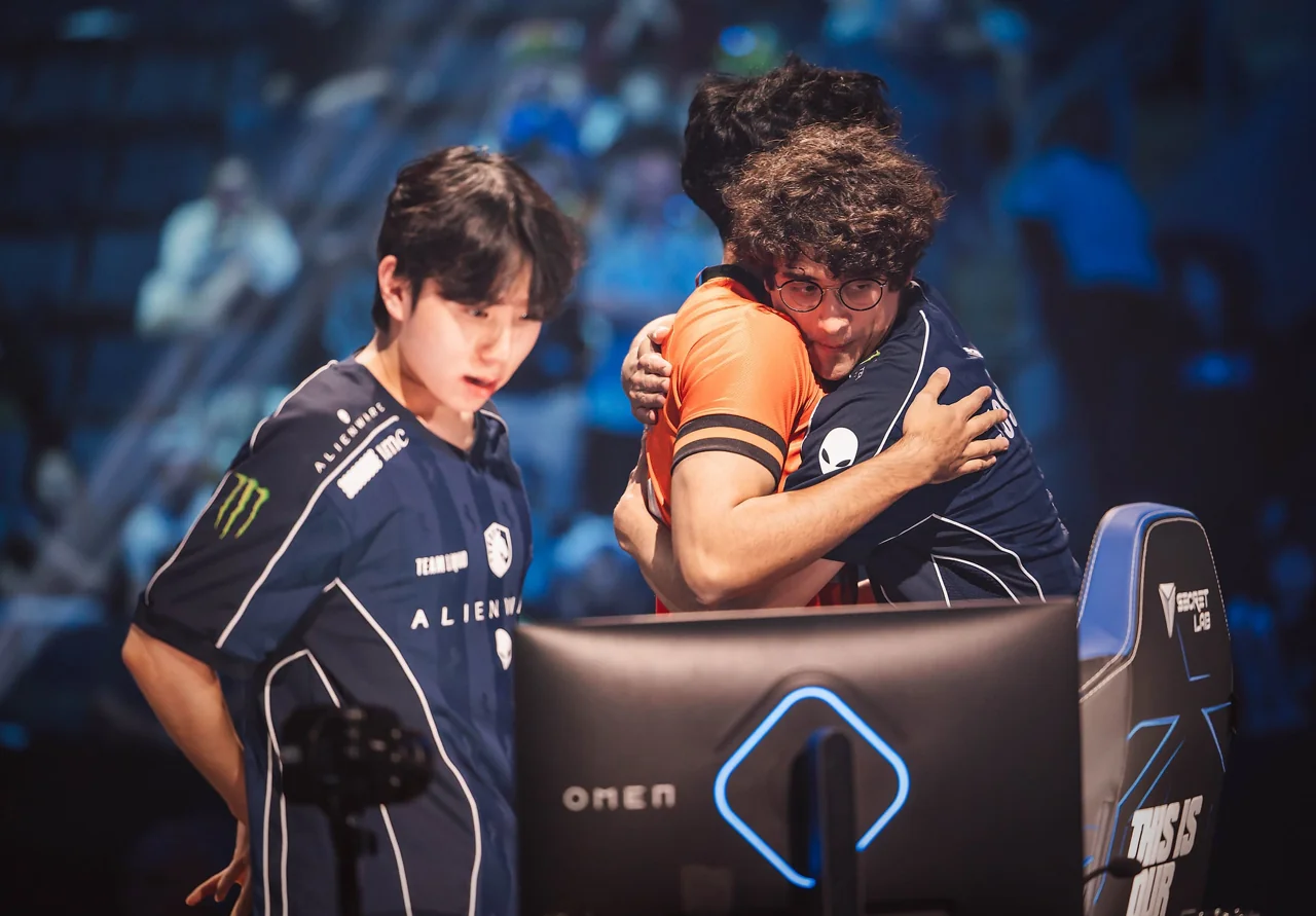 Два игрока в форме Team Liquid на турнирной сцене, один обнимает человека в оранжевой футболке